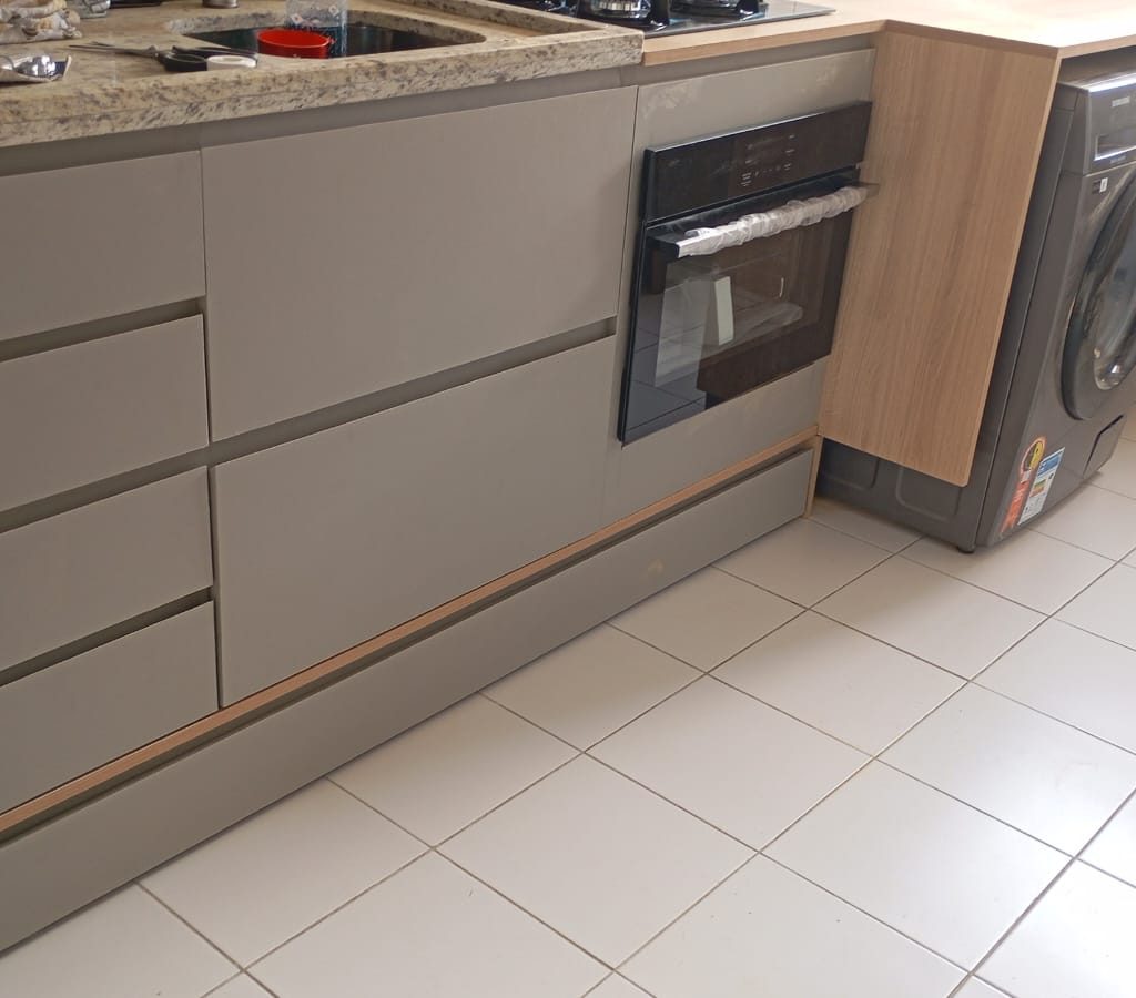 Balcão de cozinha planejada em formato L com gavetas, forno elétrico embutido e máquina de lavar roupa integrada. Projeto em MDF cinza e acabamento amadeirado.